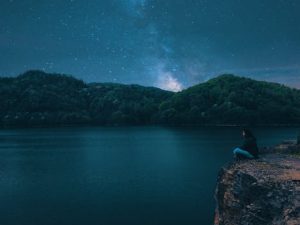 A person sits on a cliff overlooking a serene lake under a starry night sky.