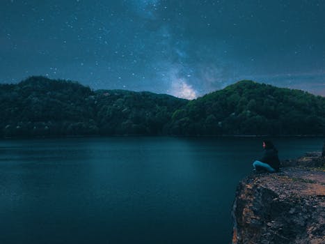 A person sits on a cliff overlooking a serene lake under a starry night sky.